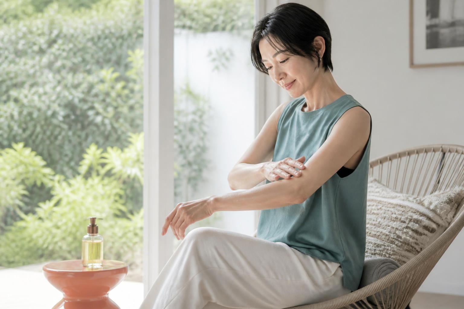 明るい窓辺で腕にボディオイルをなじませる青緑の服の女性