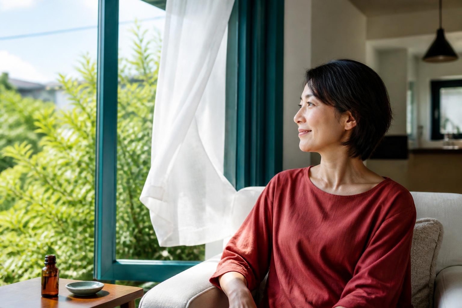 青緑の窓枠と白いカーテンのそばで、赤い服の女性が外の緑を眺める休息の時間